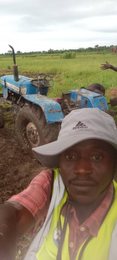 Man taking a selfie in a muddy field with a blue tractor stuck in the background.